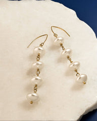 A pair of gold dangle earrings with irregular white freshwater pearls, displayed on a white textured stone slab against a dark blue background.