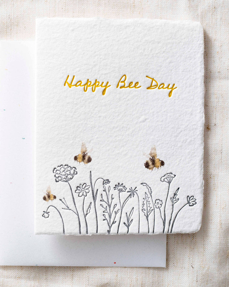 Greeting card with 'Happy Bee Day' text and bee illustrations on a textured fabric background