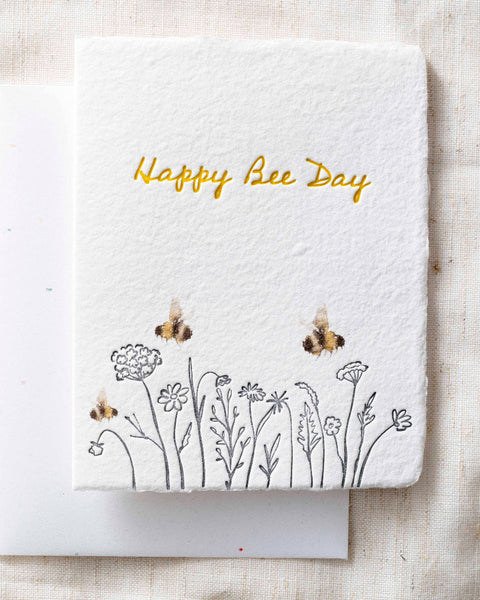 Greeting card with 'Happy Bee Day' text and bee illustrations on a textured fabric background