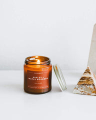 An amber glass jar candle with its lid off, showing a lit flame, on a white surface next to a geometric stone object. The label reads 'APPLES & MAPLE BOURBON'.