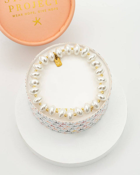 A pearl-bead bracelet with a gold star charm and a multicolor mermaid-scale patterned band, displayed on a white round pedestal next to its peach-colored branded box lid.