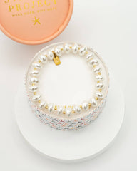 A pearl-bead bracelet with a gold star charm and a multicolor mermaid-scale patterned band, displayed on a white round pedestal next to its peach-colored branded box lid.
