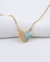 A gold heart pendant necklace resting on a white cylindrical display block. The heart is split down the middle, with one half in polished gold and the other half featuring a light blue and white marbled stone inlay.