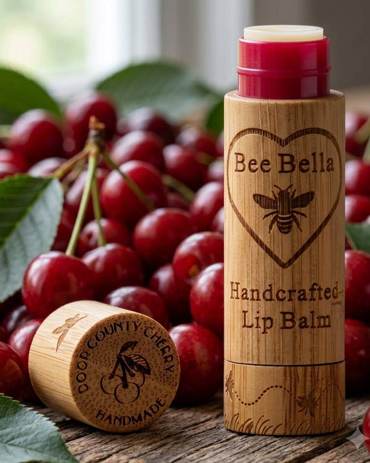 Bamboo lip balm tube labeled Bee Bella Handcrafted Lip Balm with a cherry-themed cap, surrounded by fresh red cherries and green leaves.