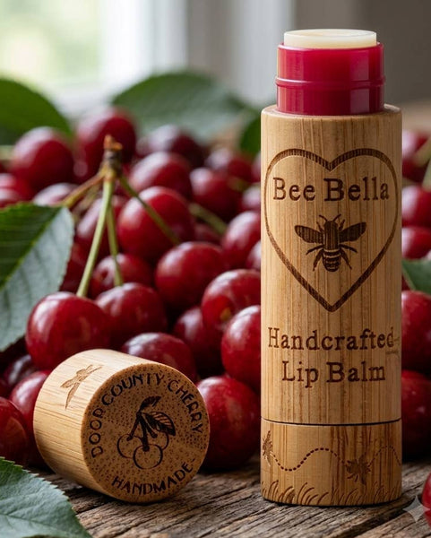 Bamboo lip balm tube labeled Bee Bella Handcrafted Lip Balm with a cherry-themed cap, surrounded by fresh red cherries and green leaves.
