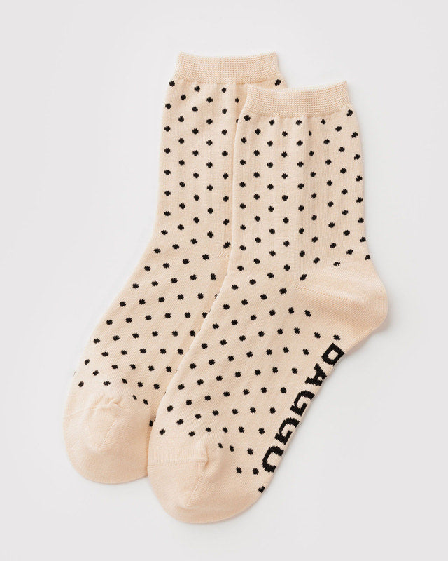 A pair of light beige crew socks with a black polka dot pattern, displayed on a clean light gray background. The word 'BAGGU' is printed in black on the side of one sock.