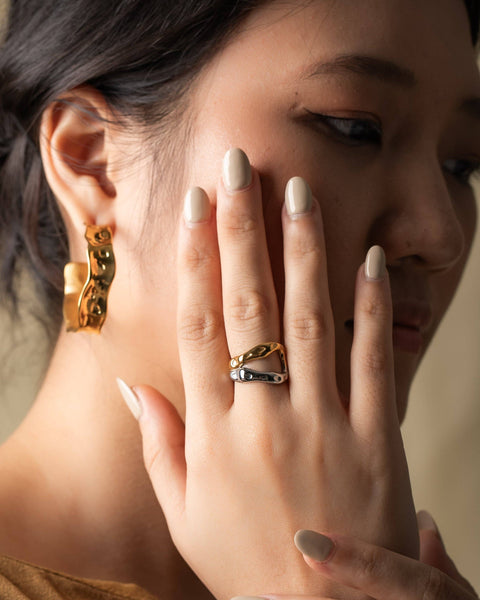 A close-up lifestyle shot of a woman wearing a gold and silver safety-pin style ring on her finger, with a matching gold huggie hoop earring on her other ear. She has light beige manicured nails and is wearing a brown top against a soft beige background.