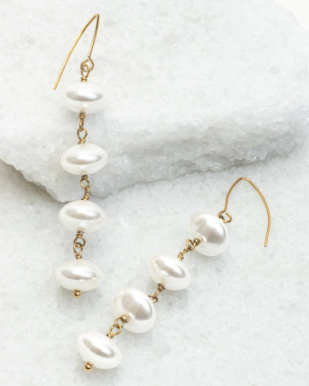 Gold-tone drop earrings with irregular white freshwater pearls, displayed on a textured white stone surface.