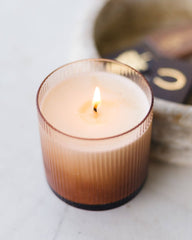 A lit candle in a ribbed amber glass jar sits on a light marble surface with a softly blurred decorative tray in the background.