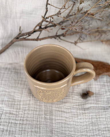 A beige ceramic mug with a handle and embossed floral detailing, placed on a textured light gray fabric surface with dried branches in the background.