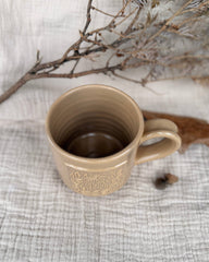 A beige ceramic mug with a handle and embossed floral detailing, placed on a textured light gray fabric surface with dried branches in the background.