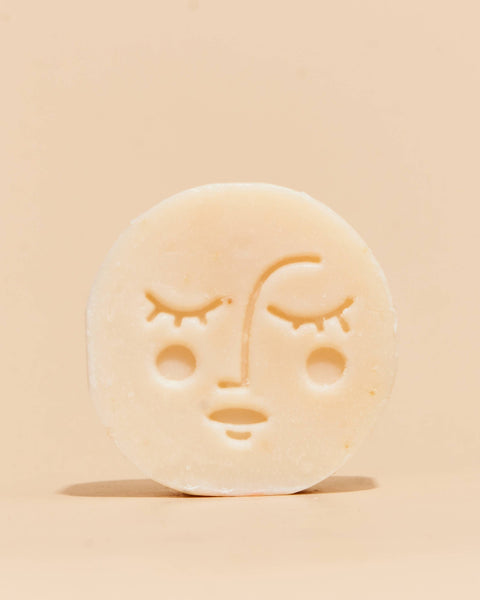 A round, off-white soap bar with a carved moon face design, standing upright on a matching beige background.