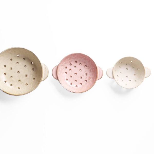 Three ceramic colanders in beige, pink, and white on a white background