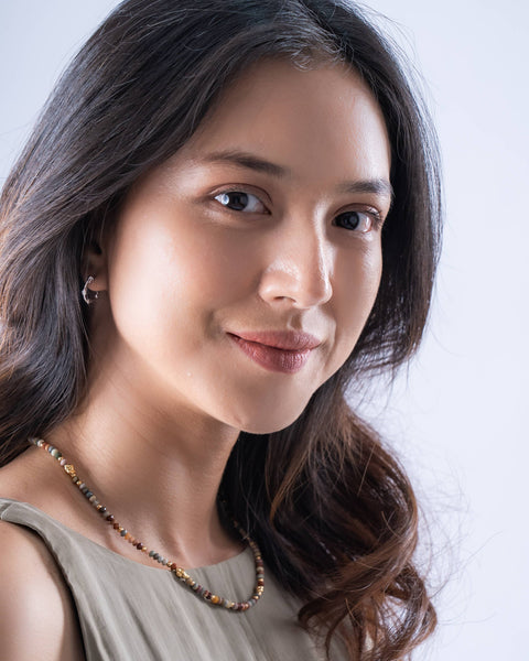 A close-up portrait of a woman with long dark brown hair, wearing a light beige sleeveless top and a beaded necklace, set against a clean light gray background.
