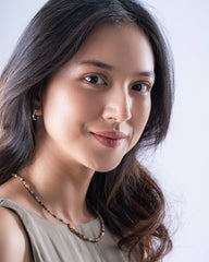A close-up portrait of a woman with long dark brown hair, wearing a light beige sleeveless top and a beaded necklace, set against a clean light gray background.