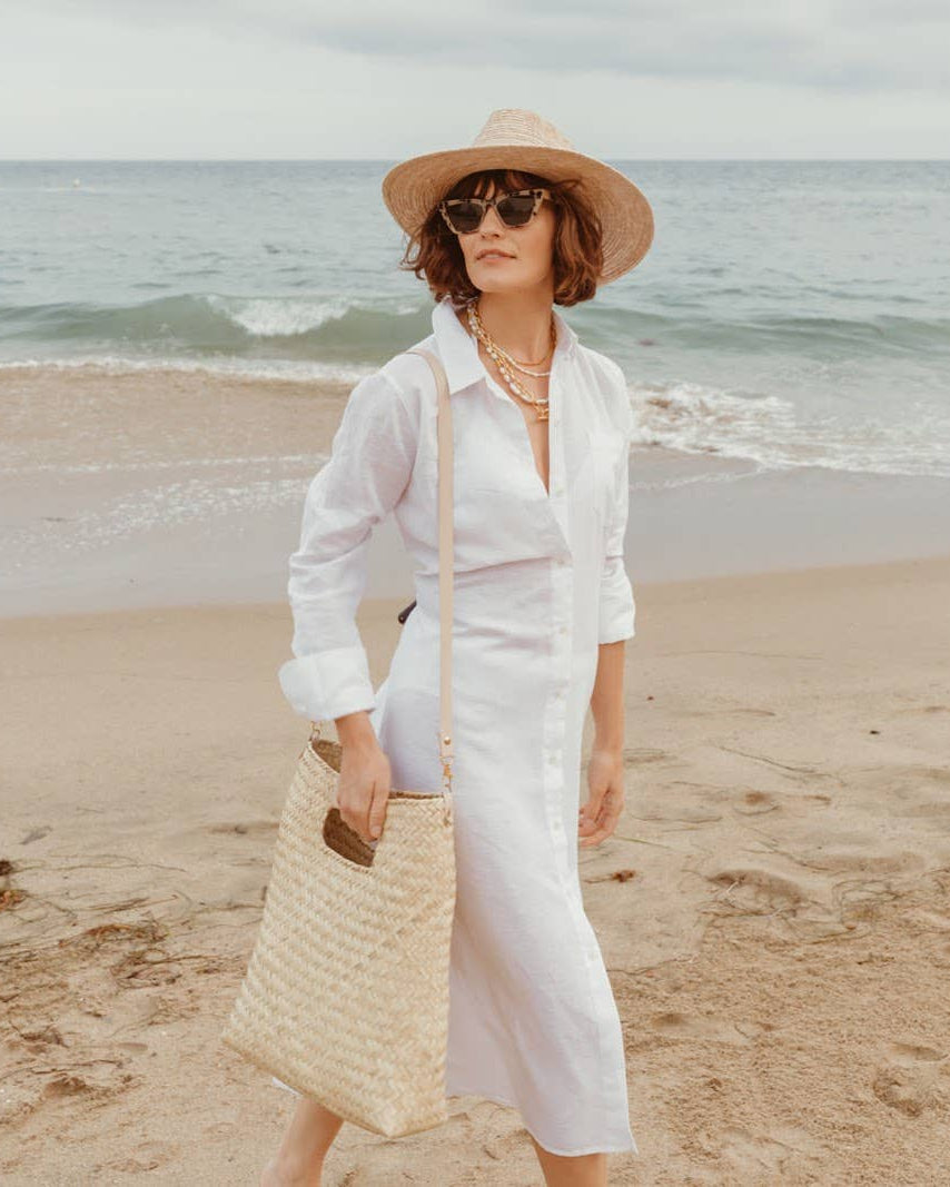 A woman stands on a sandy beach wearing a white long-sleeve midi shirt dress, a straw sun hat, sunglasses, and carrying a large woven straw shoulder bag.