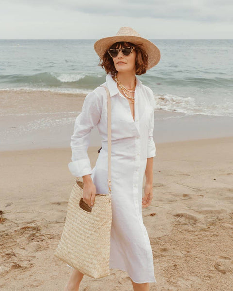 A woman stands on a sandy beach wearing a white long-sleeve midi shirt dress, a straw sun hat, sunglasses, and carrying a large woven straw shoulder bag.