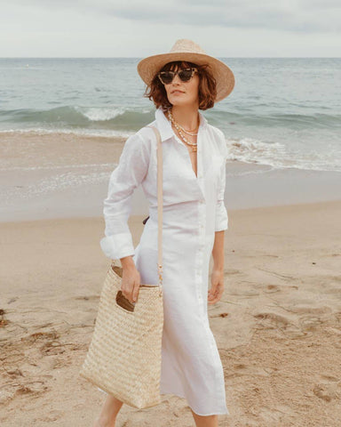 A woman stands on a sandy beach wearing a long white button-up shirt dress, a beige wide-brim hat, and holding a large woven straw tote bag. The ocean and cloudy sky are visible in the background.