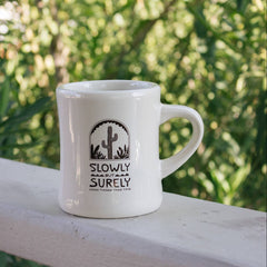 White mug with 'Slowly Surely' design on a wooden railing with greenery in the background