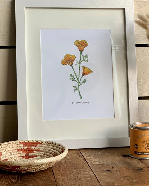 Framed botanical print of flowers on a wooden surface with a vase and candle.