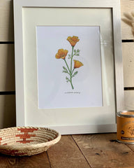 Framed botanical print of flowers on a wooden surface with a vase and candle.