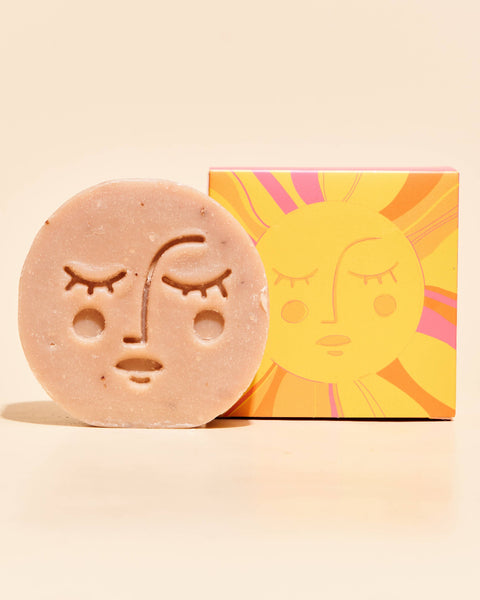 A round, textured natural soap bar with an engraved face sits in front of its colorful packaging box, which features a stylized sun graphic, on a warm cream background.