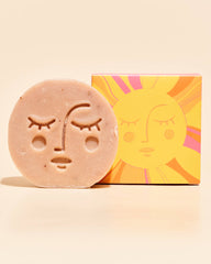 A round, textured natural soap bar with an engraved face sits in front of its colorful packaging box, which features a stylized sun graphic, on a warm cream background.