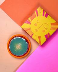 A square product box with a yellow sun illustration sits next to an open round jar with a teal lid and orange rim on a pastel color-blocked background.