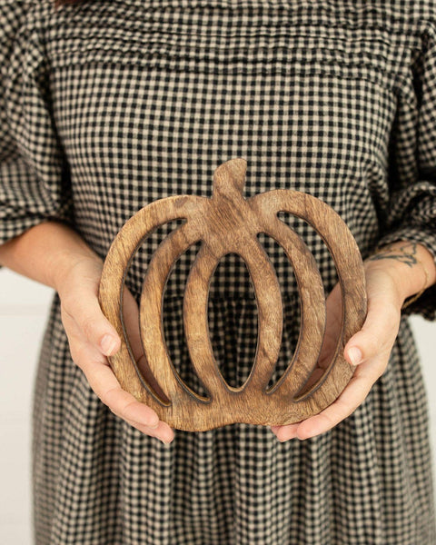 A person in a black and white gingham dress holds a carved wooden pumpkin decoration in both hands against a plain light background.