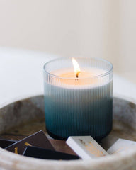 A lit candle in a ribbed clear glass jar with a blue-to-white ombre design, styled in a round stone tray with small cards on a light surface.