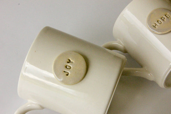 Two white mugs with 'HOPE' and 'JOY' on a light gray background