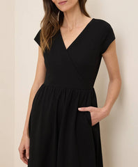 A woman wearing a black wrap-style dress with cap sleeves, a V-neckline, and a gathered waist with side pockets, photographed against a plain light beige wall.