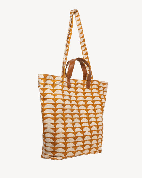 A canvas tote bag featuring a repeating ochre orange and cream semicircle pattern, with solid brown leather handles and trim, shown against a plain light gray background.