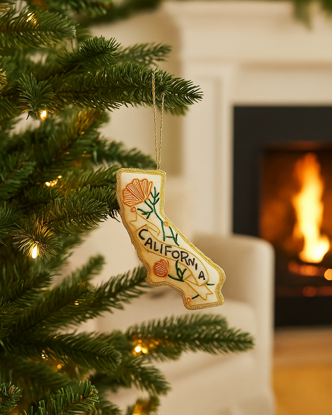 A California-shaped Christmas ornament hanging from a decorated Christmas tree in front of a cozy fireplace with a burning fire visible in the background.