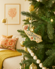A close-up of a decorated Christmas tree in a cozy living room, featuring a California-shaped ornament, gold baubles, white pom-pom garlands, and warm ambient lighting.