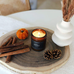 Candle labeled 'Home Holiday' on a wooden tray with cinnamon sticks, pinecones, and a small pumpkin.