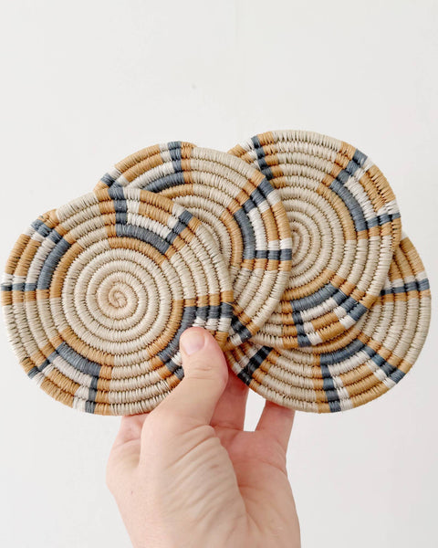 Two round woven coasters with spiral patterns in beige, brown, and gray, held by a hand against a plain white background.