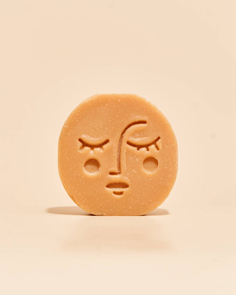 A single round, light tan soap bar with an embossed sad face, standing upright on a matching light beige background.