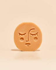 A single round, light tan soap bar with an embossed sad face, standing upright on a matching light beige background.
