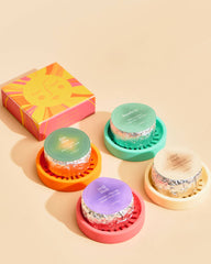 Four small round packaged items arranged on colorful circular stands next to a bright pink and yellow box on a warm beige background.
