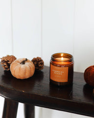 A lit amber glass jar candle labeled 'Pumpkin Spice' sits on a dark wooden stool, styled with small pumpkins, pinecones, and cinnamon sticks against a white paneled wall.