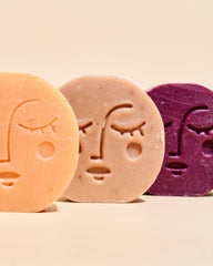 Four round handmade soaps with embossed face designs in cream, peach, taupe, and purple, arranged in a row on a light beige background.