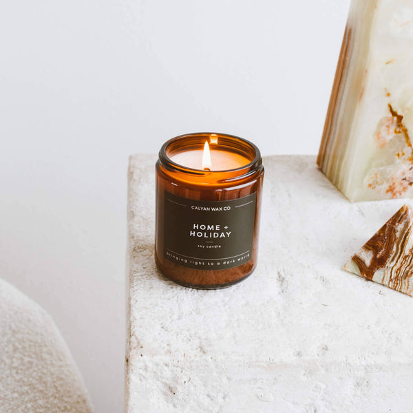 Candle in an amber glass jar labeled 'Home + Holiday' by Calyan Wax Co on a white surface.
