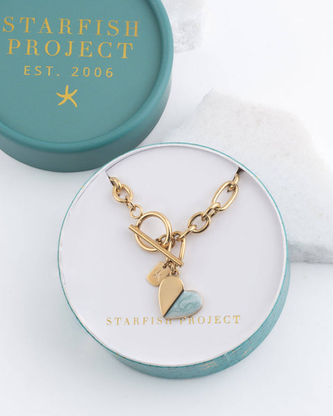 A gold-tone charm bracelet with heart charms, displayed inside an open round gift box. The box lid, which is teal with gold lettering reading STARFISH PROJECT 