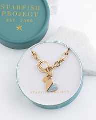 A gold-tone charm bracelet with heart charms, displayed inside an open round gift box. The box lid, which is teal with gold lettering reading STARFISH PROJECT 