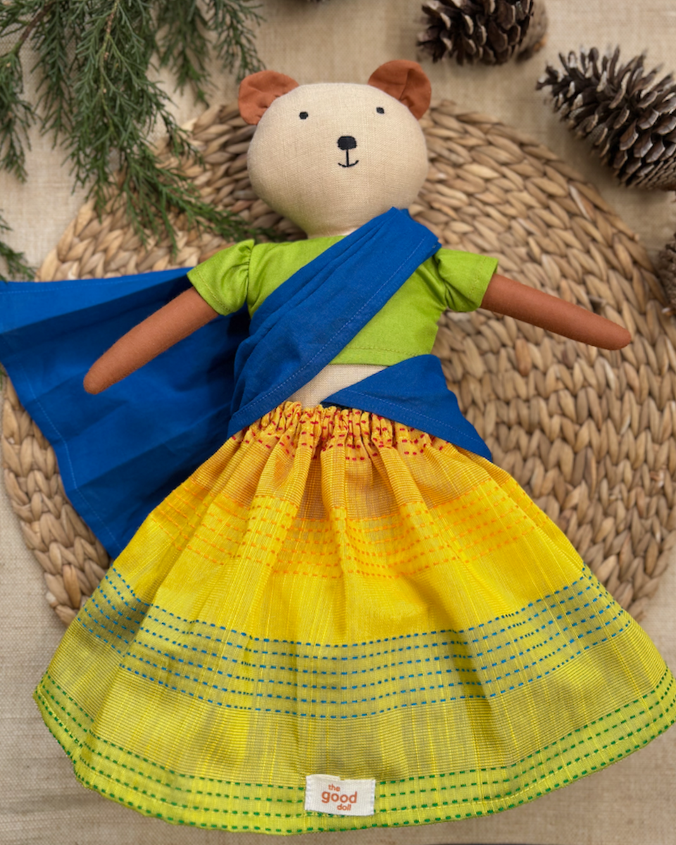 Toy bear wearing a blue and yellow dress on a woven mat with pine cones and branches.