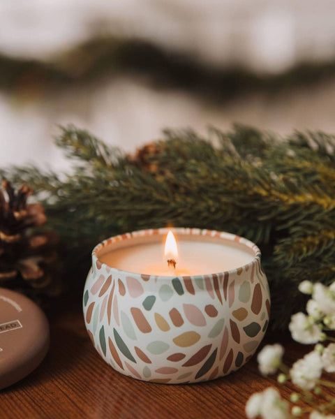 A lit candle in a white ceramic holder with a muted multicolor leaf pattern, styled on a wooden surface with pine branches, a match jar, and small white flowers.