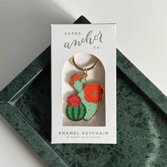 Cactus-shaped enamel keychain in a Paper Anchor Co. packaging on a green marble surface.