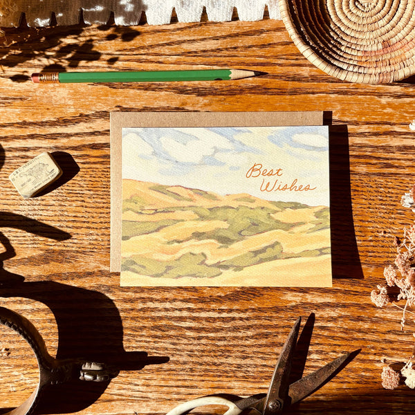 Note card with landscape design and 'Best Wishes' text on a wooden surface with tools and a woven basket.