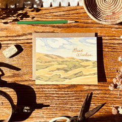 Note card with landscape design and 'Best Wishes' text on a wooden surface with tools and a woven basket.